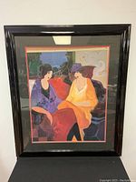Full view of framed art print by Tarkay showing two seated women with colorful stylized forms, framed in black glossy frame with matting.
