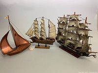 Three model ships displayed in a row on a neutral background, showing copper sailboat and two wooden ship replicas.