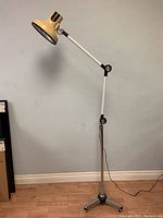 Full view of vintage adjustable industrial floor lamp standing approx 72 inches tall, beige shade, white adjustable arm, metal base on casters on wood floor against light blue wall.