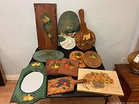 Wide view of eleven wooden wall art pieces and cheeseboards painted with fruit, floral, and village scenes arranged on black table.