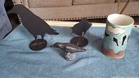 Front view of two black metal raven sculptures, small clay raven figurine, and painted clay cup/vase on a blue cloth background.