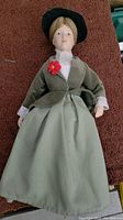 Full body view of the porcelain doll showing the green jacket, white lace blouse, green skirt, and red flower brooch.