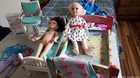 Two dolls—one dressed and one unclothed—lying on two different doll beds with blankets and headboards, showing bedding and bedframes.
