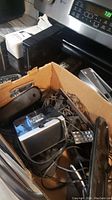 Box containing various electronics including remote controls, cordless phones, and assorted cords tangled together.