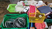 Large green plastic jug with a lid and handle, assorted doilies, new tablecloths and tea towels, yellow wire storage rack, and blue rug alongside bin of pots and pans shown in the background.