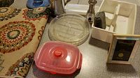 Assorted kitchen and household items including a medallion patterned fabric mat, insulated carafe, red plastic food container with lid, salt shaker, cleaning caddy with sponge, sewing thread container, and picture frame with mat.