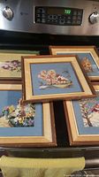 Five framed vintage needlepoint artworks laid out showing landscapes with trees, water, and houses on blue fabric