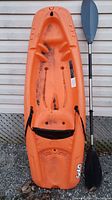 Top view of the used orange Solo kayak with mounted black seat strap, accompanied by a black and silver paddle resting next to it, showing usage marks on the kayak.