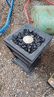 Square black outdoor fire pit with central burner and faux coals on top, placed on gravel ground with a green propane tank in background.