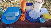 Wide shot showing all plastic dishware and storage items arranged on brown fabric outdoors, includes blue trays, orange cups and cutlery, and assorted Tupperware containers.