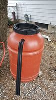 Orange cylindrical rain barrel with attached black flexible overflow hose and black lid with mesh screen, placed outdoors near a tree and house exterior.