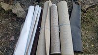 Overview of three carpeted rugs and one rolled doormat, all rolled and laid out side by side outdoors on grass and gravel