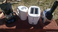 Overview of all four small appliances arranged outdoors on a brown towel: coffee maker, white kettle, white toaster, and Yonanas black and silver soft serve machine.