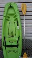 Photo showing a green sit-in kayak for kids with a black adjustable strap seat and a yellow blade paddle lying on the ground next to it.