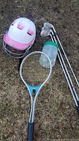Photo showing all items on grass: batting helmet, tennis racket, three golf clubs, container with golf tees.