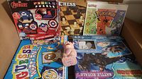Box containing seven assorted kids games: Avengers pixel art puzzle, chess, Pokemon puzzle, Guess Who, Battle Lego and Battle Royale games, and a pig plastic toy