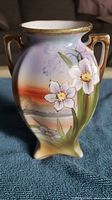 Front view of vintage Nippon hand painted mini vase showing raised white flowers and scenic background with two gold-accented handles.