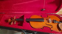 Full view of violin inside the red velvet-lined case showing violin body, fingerboard, tailpiece and some strings.