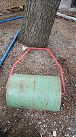 Green metal cylindrical yard roller with red handle leaning against tree outside, showing signs of rust and missing plug