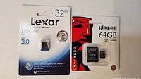 Packaged Lexar 32GB USB 3.0 flash drive alongside Kingston 64GB microSD card with SD adapter, unopened packaging visible.