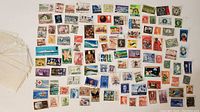 Photo shows a broad assortment of loose postage stamps in varying colors and sizes spread out with some postal marks visible.