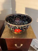 Angled front view of cloisonne bowl showing black enamel exterior with red floral motifs on brass-tipped surface