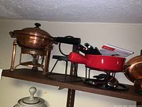 Photo showing a shelf with three fondue sets, including a brass set on the left, a red ceramic set in the middle with forks and ladle, and a partially visible brass item on the right.
