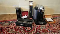 Photo of all items including the coffee maker, two milk frothers, three boxes of capsules and accessories on a rug.