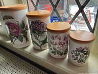 All 4 Portmeirion Botanic Garden ceramic canisters with wooden lids arranged in a row, showing varied sizes and floral artwork on containers.