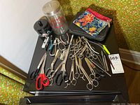 Overview of large assortment of scissors, nail clippers, tweezers, and vintage containers on small black table.