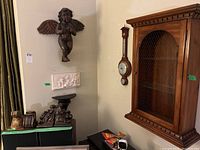 Wide view showing carved wood cherub/angel wall hanging, plaster relief plaque mounted under it, vintage barometer to the right, and large wooden display cabinet with chicken wire on the right wall.