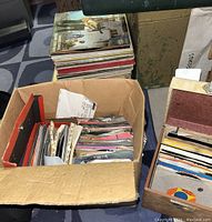 Top-down view of a cardboard box filled with many 45 RPM vinyl records in paper sleeves, stacked vertically inside.