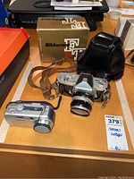Photo showing both cameras on a wooden table along with Nikkormat box and leather camera case, illustrating the complete lot contents.