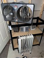 Overview showing dual Pelonis fan unit, white DeLonghi oil-filled heater, and smaller heaters on shelf in basement.