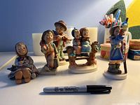 Front view of five Goebel Hummel figurines displayed on white surface with a Sharpie for scale.