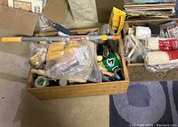 Photo showing wooden crate filled with various paint supplies including a long extension pole, rollers, brushes, and more.