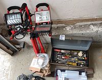 Two Commercial Electric work lights on red tripod stands, metal toolbox partially open showing hand tools, pewter towel holder, and white Delta soap dispenser packed