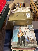Photo showing a gold carrying case with a box and stack of vinyl LP records including an album featuring musicians holding guitars, text in Hebrew.
