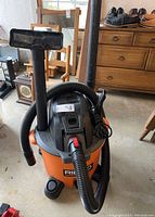 Orange Ridgid shop vacuum with hose coiled on top and attached to floor nozzle, black lid with power cord wrapped around it, standing on concrete floor with wooden furniture in background.