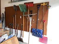 Garage wall mounted peg board with various garden tools hanging, including shovels, roof rake, pitchfork, root feeders, ax, broom, and scrub brush.