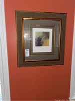 Framed signed wall art hung on a burnt orange wall, showing overall condition and appearance.