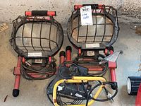 Photo showing two large Husky work lights with red frames and metal protective cages side by side with a smaller black and yellow work light on the floor.