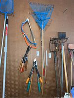 Photo of hanging garden tools including two blue metal leaf rakes, two bow saws with colored handles, hand clippers, and metal hand rake.
