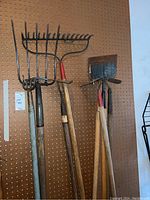 Photo of yard tools with metal rake, spading fork, garden hoe, and other wooden handled tools hanging on pegboard, showing condition and variety.