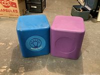 Two cube-shaped stools side by side on concrete floor, blue and purple.