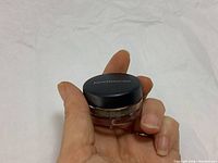 Hand holding mini jar with black lid labeled Bare Minerals on a white crumpled background