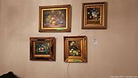 Four framed art pieces on wall displaying floral and fruit still life paintings with ornate gold frames.