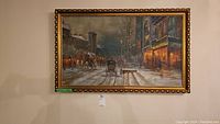 Close front view of first framed cityscape painting showing street scene with horse carriage, pedestrians, and lit storefronts in the evening or night, mounted in ornate gold frame.