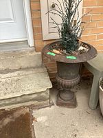 Cast iron urn-shaped planter positioned near concrete steps with brick wall backdrop. Weathered surface with natural debris inside.
