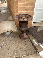 Photo of one weathered cast iron planter with urn shape, decorative edging, and square base placed on an outdoor concrete surface near a brick wall.